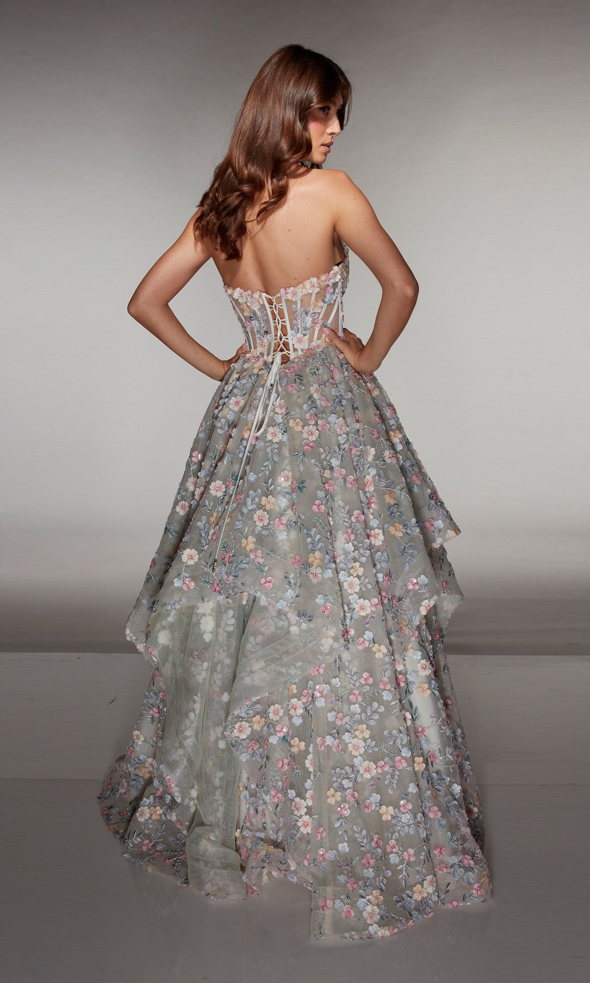 A woman with long brown hair faces away, wearing the Alyce 62048 long prom dress with a strapless corset bodice and a tiered, flowing skirt embellished with delicate floral appliqué.